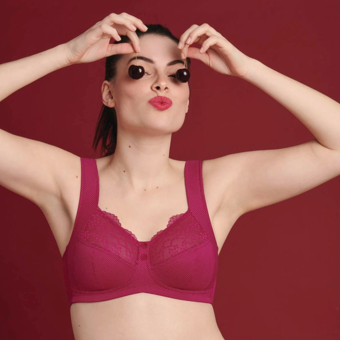 A woman wearing a cherry red, wirelessly supported, knitted fabric bra with French polka dot charm, posing with one hand raised to her head.