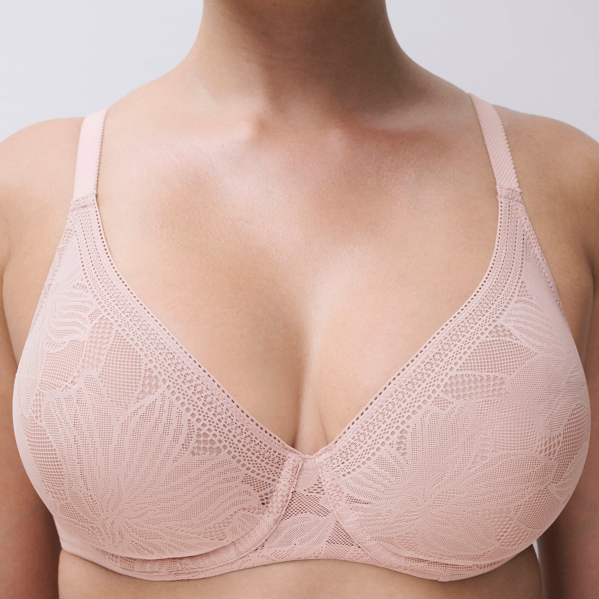 A close-up of a model wearing a light pink floral lace underwire bra against a neutral light gray background.