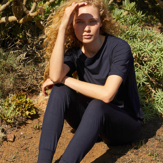 A person with curly hair sitting outdoors wearing a dark short sleeve shirt and pants.