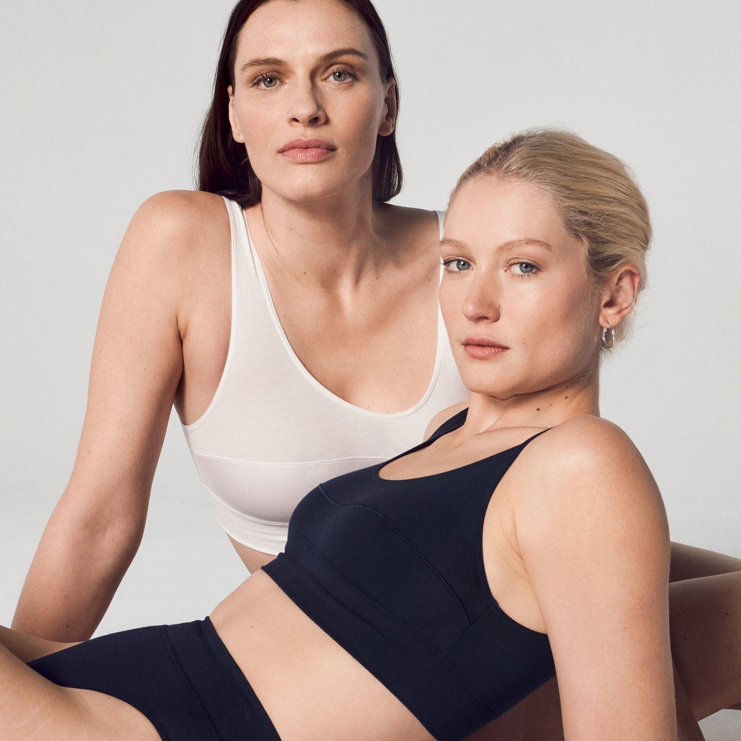 Two models, one in a white and one in a black bralette, posing on a neutral background.
