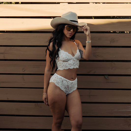 Woman in a white lace outfit and cowboy hat standing against a wooden wall.
