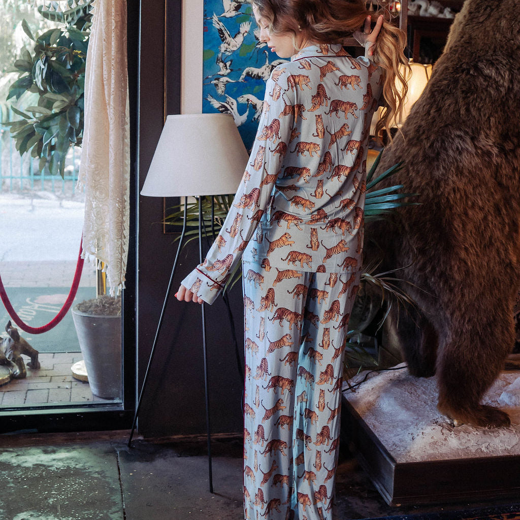 A woman stands indoors wearing a matching pajama set with an allover fox print. The set includes a long-sleeve top and a long pair of pants in a light gray base with orange, brown, and black foxes.