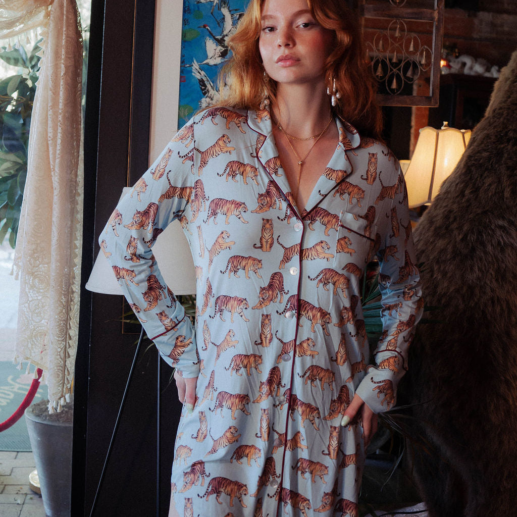 A woman wearing a light blue long-sleeve pajama shirt with an all-over orange and brown tiger print, standing in a rustic room with a framed painting and warm ambient lighting.