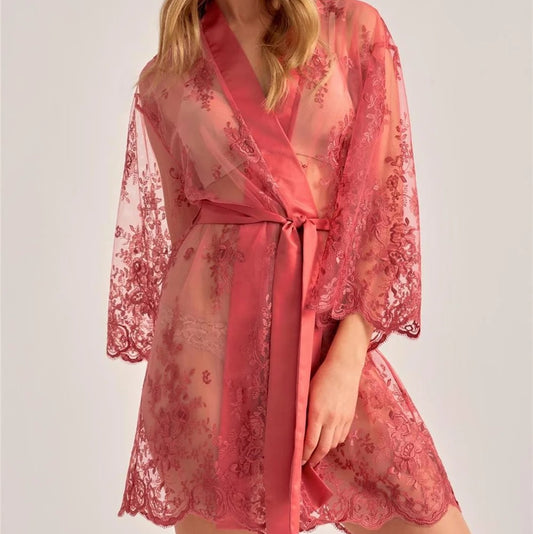 A woman modeling a sheer pink lace robe with a matching satin tie belt, photographed against a plain light gray studio background.