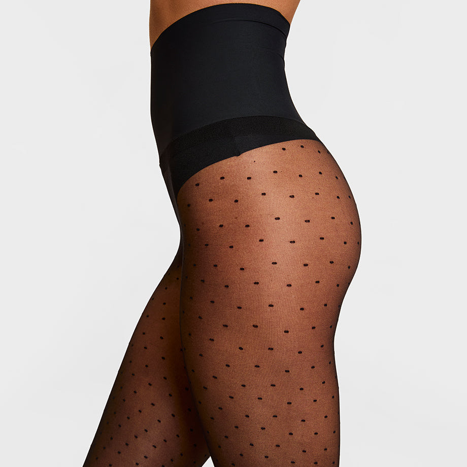 Close-up side view of a model wearing sheer black polka dot tights with a wide black waistband, shown against a light gray studio background.