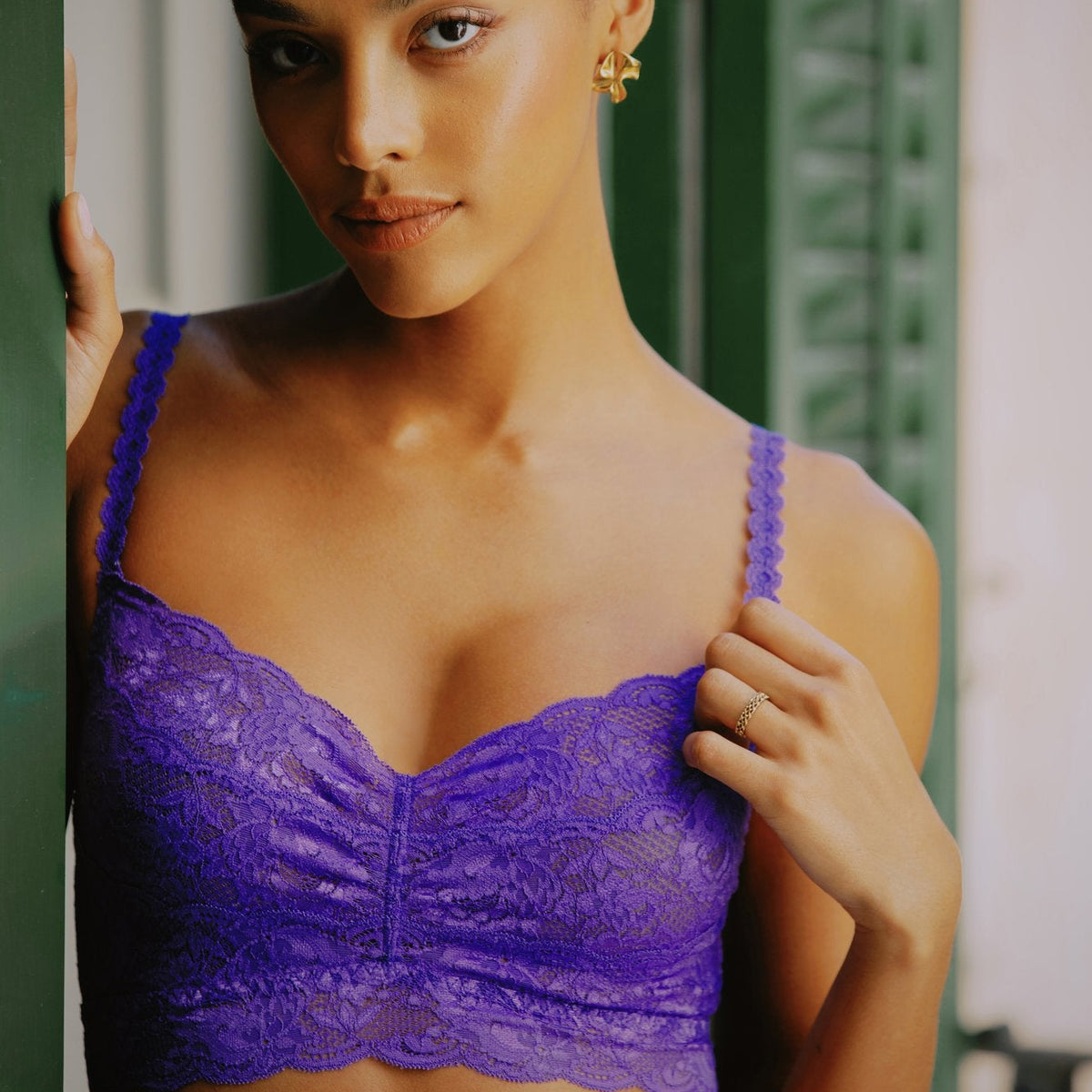 A woman wearing a purple lace underwire bra, posing against a green shuttered window background.