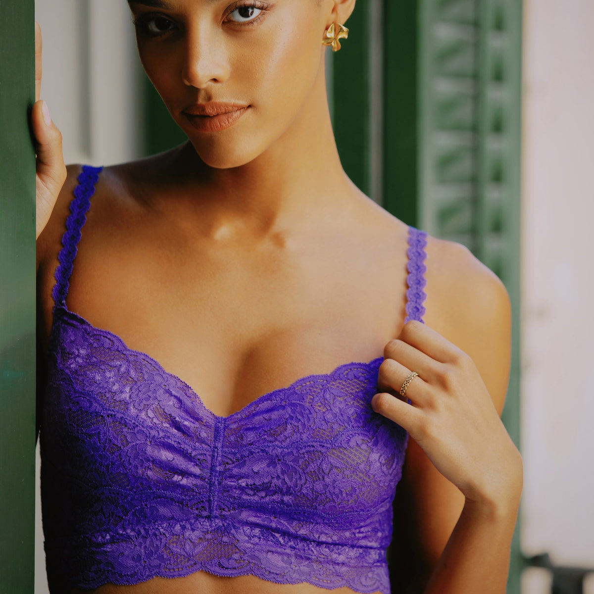 A woman wearing a purple lace underwire bra, posing against a green shuttered window background.