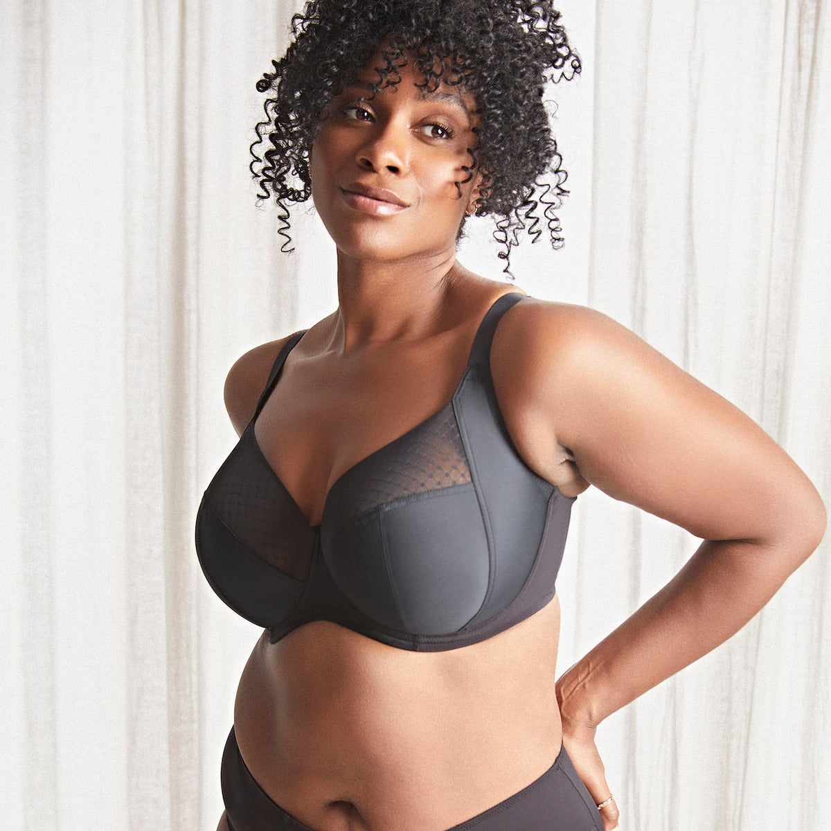 Woman wearing a dark gray bra and underwear set against a white curtain background