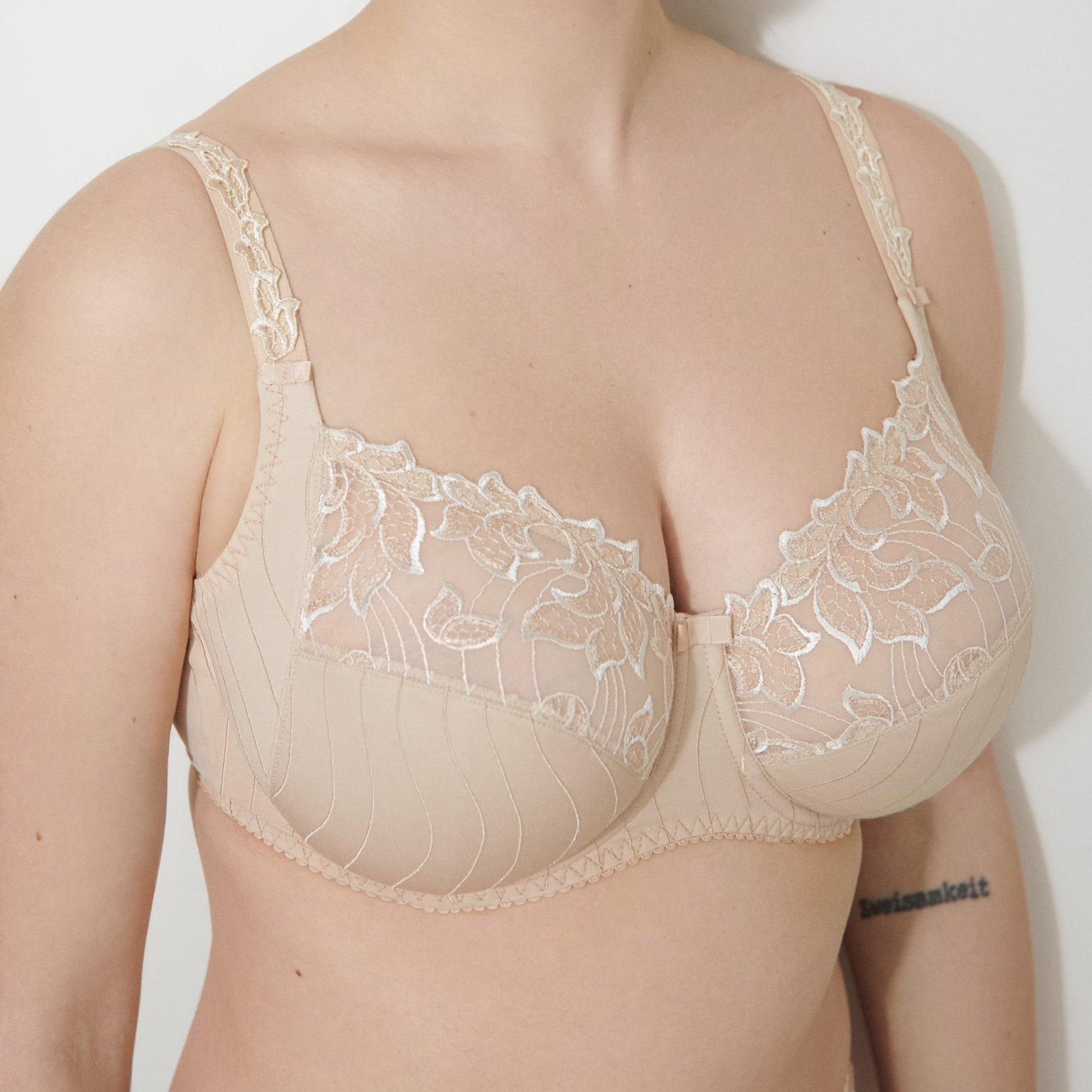 A woman wearing a beige full cup bra with embroidered detailing on the cups and straps.
