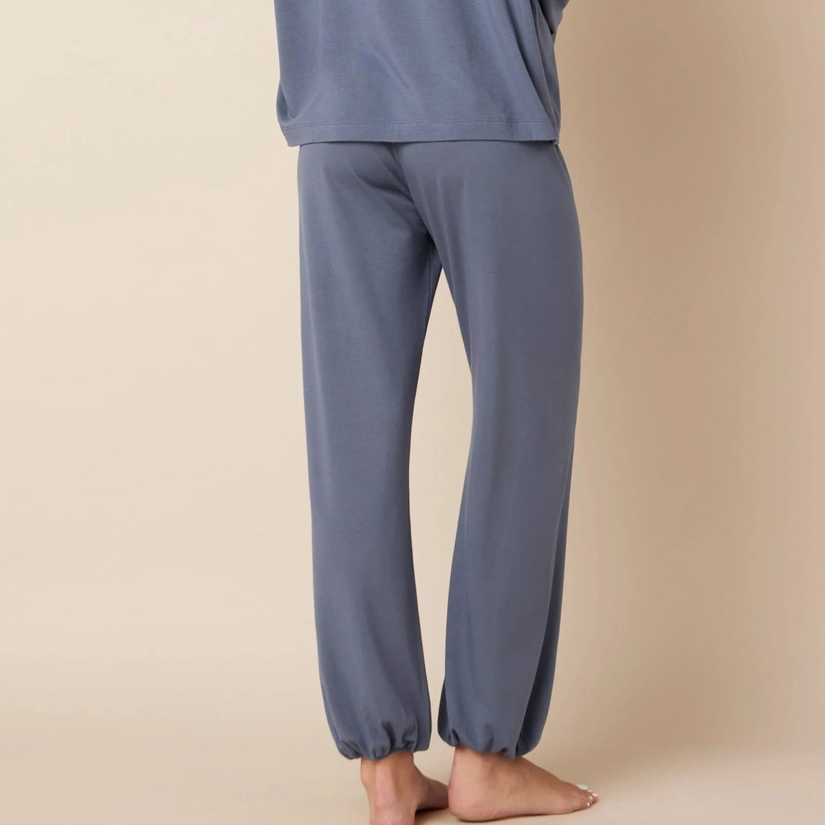 A woman stands barefoot against a plain beige studio background, wearing a matching two-piece lounge set consisting of a loose long-sleeve top and relaxed-fit pants in a muted dusty blue-gray color.