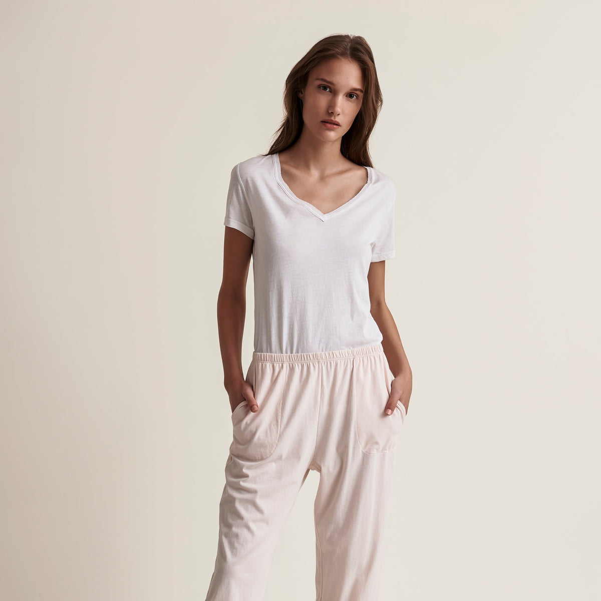 A woman modeling a two-piece loungewear set consisting of a white short-sleeve V-neck top and light blush pink cropped pants with side pockets, set against a solid warm beige studio background.
