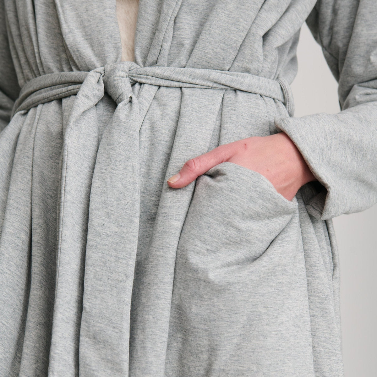 Close-up of a light gray heathered robe with a tie belt and front patch pocket, shown on a person whose hand is in the pocket.