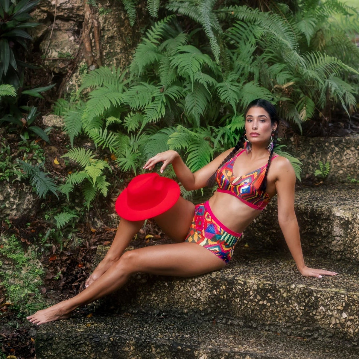 Woman in colorful outfit and red hat sitting on stone steps with greenery
