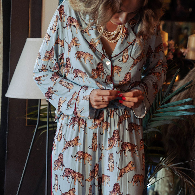A woman wearing a light blue pajama set with an all-over orange and black tiger print, standing in a dimly lit indoor setting with plants and a lamp in the background.