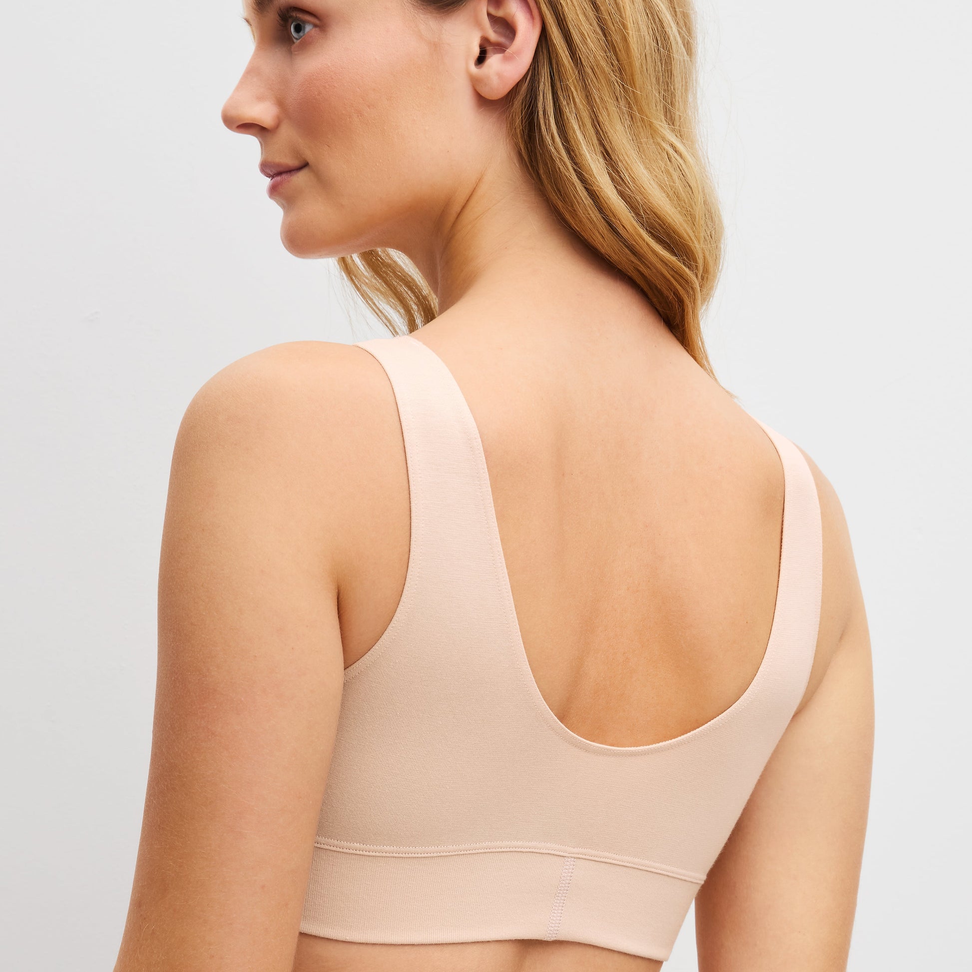 A woman shown from the back wearing a light beige/nude wireless bra with wide shoulder straps and a deep open back, photographed against a clean light gray studio background.