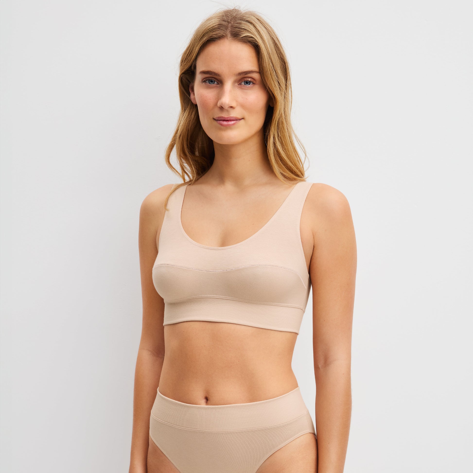 A woman modeling a matching beige two-piece underwear set consisting of a scoop-neck bralette and high-waisted briefs, photographed against a light gray studio background.