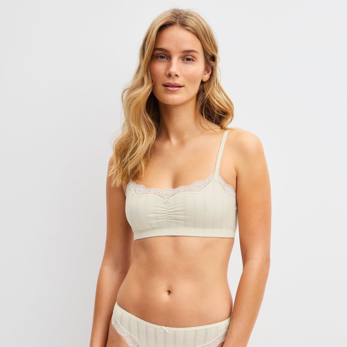 A blonde model wearing a two-piece lingerie set in a light cream color with lace trim, consisting of a bralette and matching panties, against a plain light gray background.