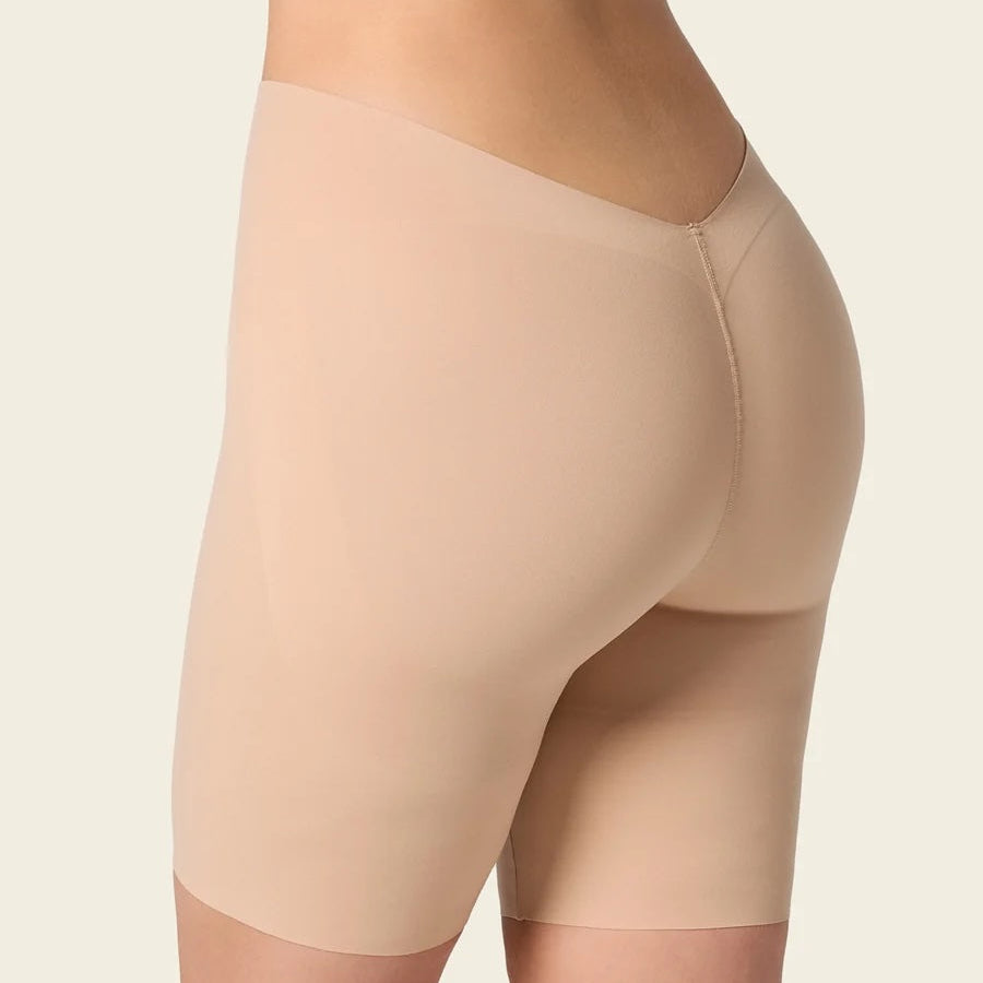 Close-up side view of a model wearing seamless shapewear briefs in a light nude beige color, showing the hip and thigh area against a plain light beige background.