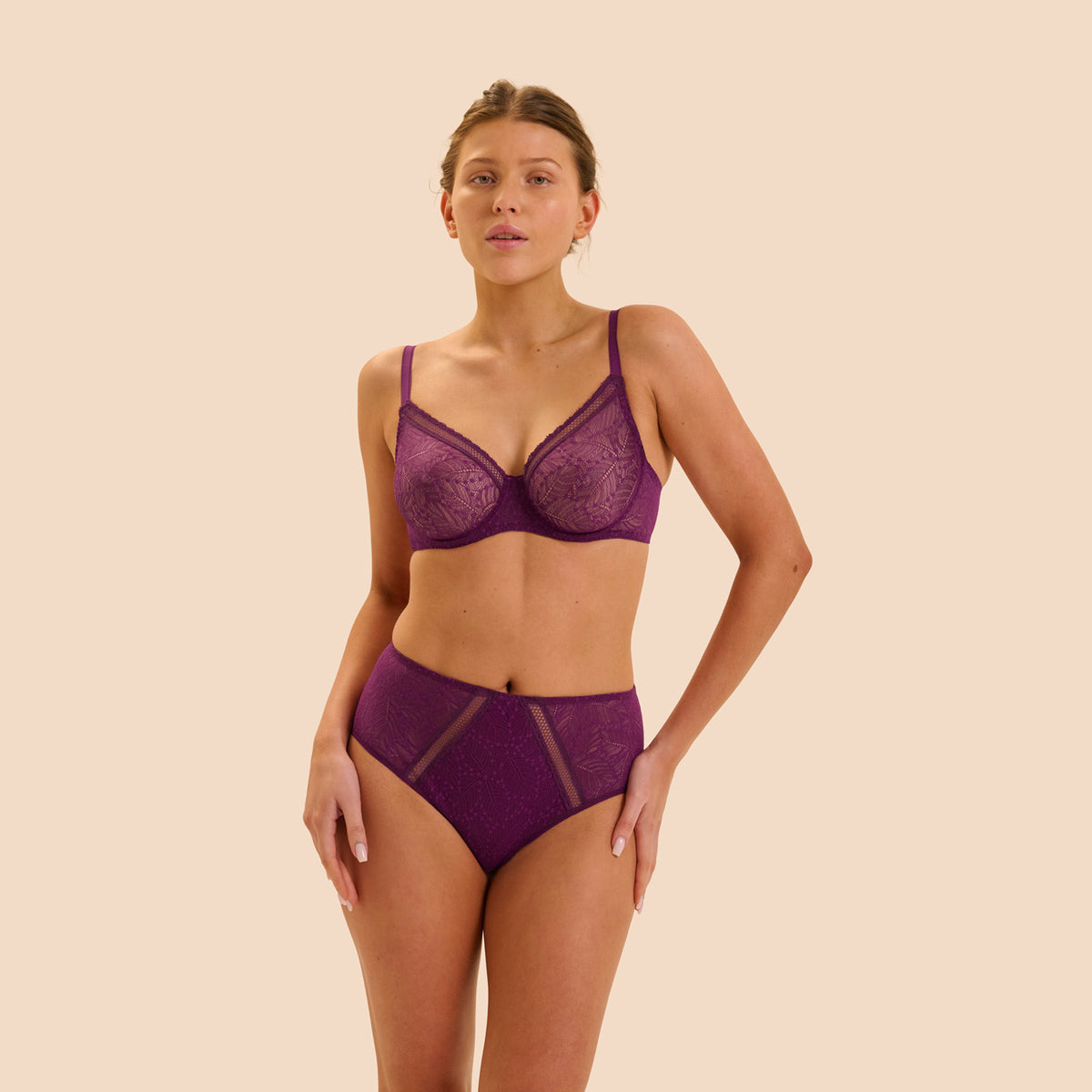 A woman modeling a matching purple lace lingerie set against a solid beige background. The set includes a sheer underwire bra and coordinating high-waisted briefs with mesh side panels.