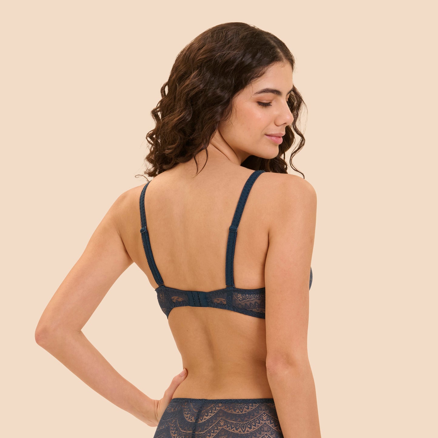 A woman shown from the back wearing a dark navy lace bralette and matching lace bottoms against a solid beige studio background.