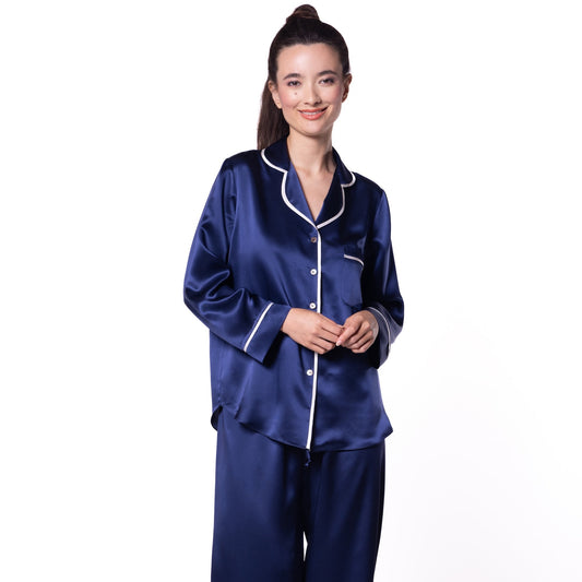 A woman modeling a two-piece navy blue satin pajama set with white piping details, consisting of a long-sleeve button-up top and matching wide-leg pants, against a plain white background.