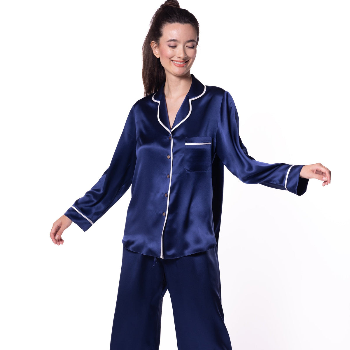 A woman modeling a two-piece navy blue satin pajama set with white piping details, consisting of a long-sleeve button-up top and matching wide-leg pants, against a plain white background.
