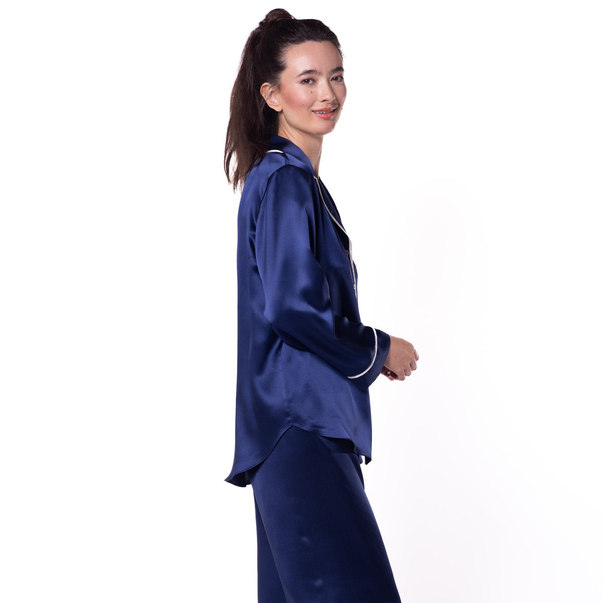 A woman modeling a navy blue satin pajama set consisting of a long-sleeve top and matching wide-leg pants, shown from a side profile against a white studio background.