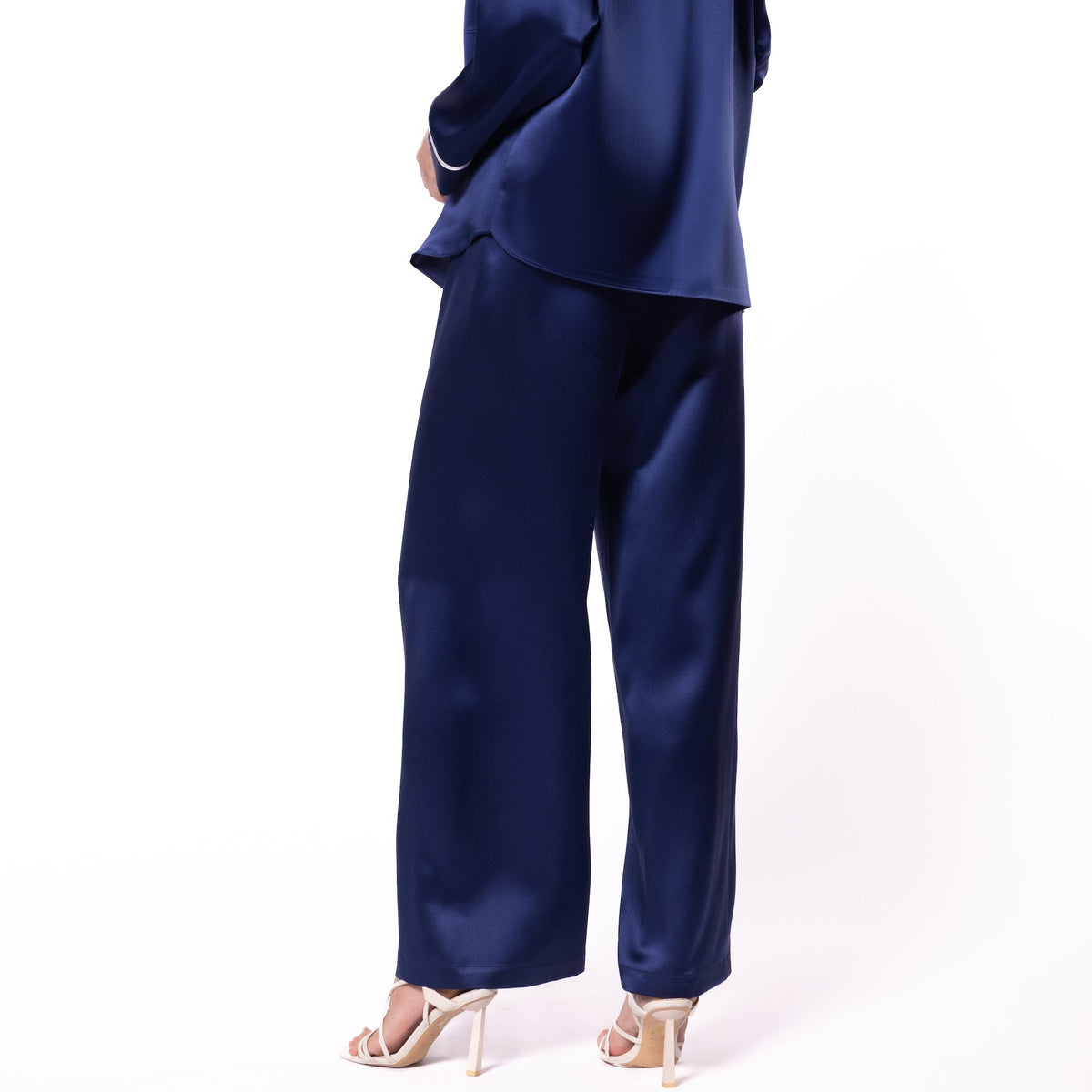 A woman modeling a navy blue satin pajama set, featuring a long-sleeve top with a high collar and matching wide-leg pants, shown from a three-quarters back view against a white background.