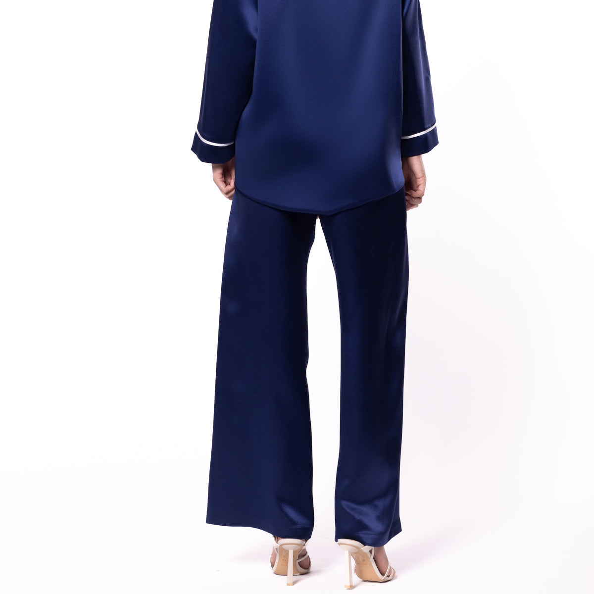 A woman viewed from the back wearing a navy blue satin pajama set with white piping trim on a plain white studio background.