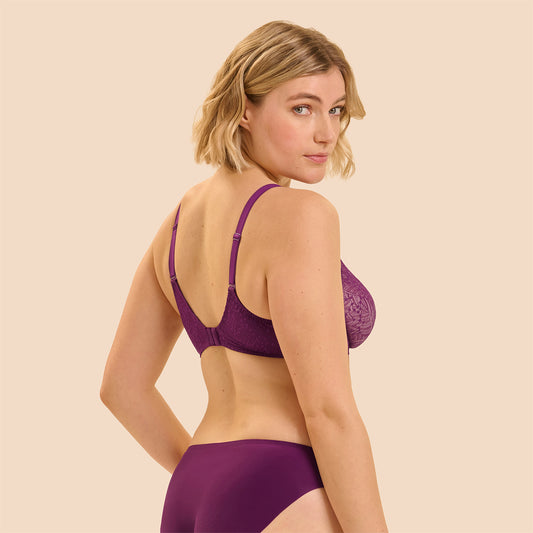 A woman modeling a purple lace bra and matching high-waisted briefs against a solid beige background, shown from a three-quarters back view.
