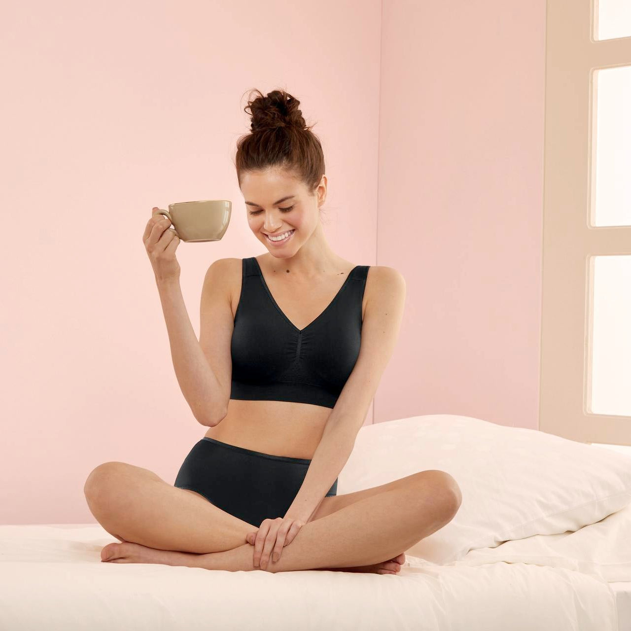 Woman in black lingerie holding a cup on a bed with pink walls