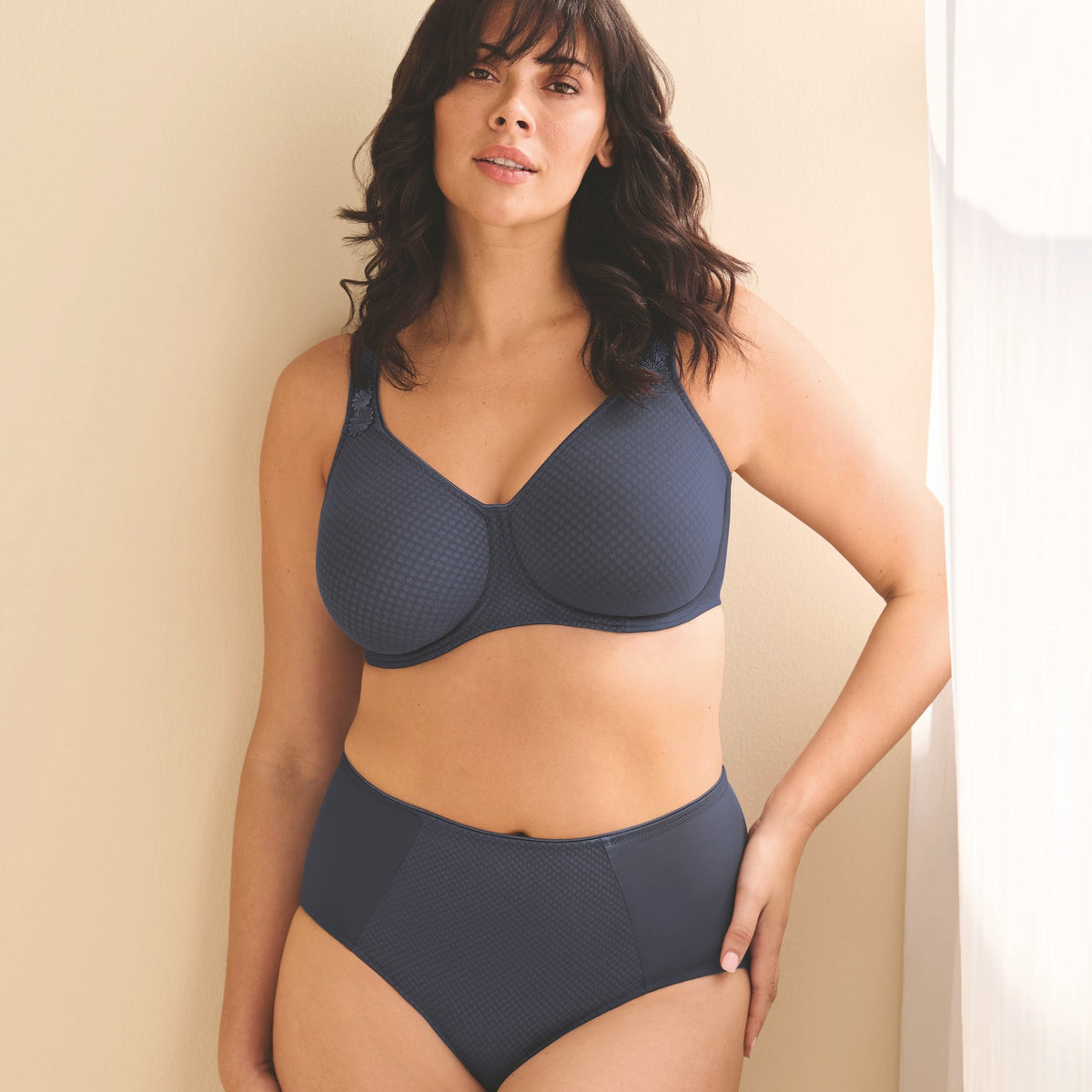 A woman modeling a dark slate blue two-piece lingerie set consisting of a textured bra and matching high-waisted briefs, standing against a warm beige wall.