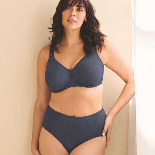 A woman modeling a dark slate blue two-piece lingerie set consisting of a textured bra and matching high-waisted briefs, standing against a warm beige wall.