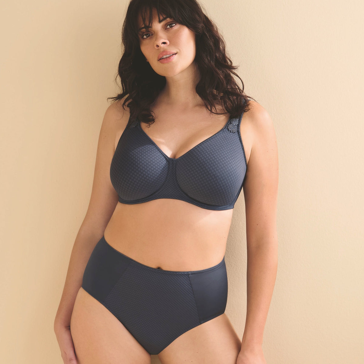 A woman modeling a dark gray two-piece lingerie set consisting of a textured underwire bra and matching high-waisted briefs, posed against a solid beige background.