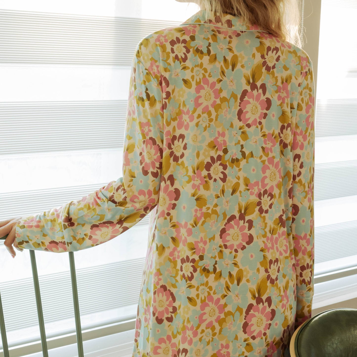 Woman wearing a floral dress standing by a window with blinds.