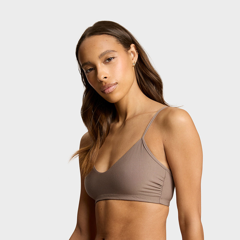 A woman modeling a matching taupe brown bralette and high-waisted brief set against a light gray studio background.