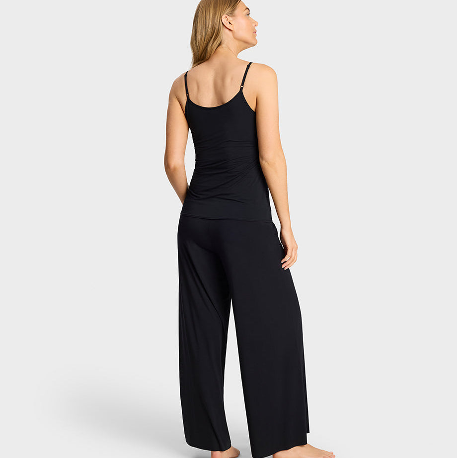 A woman stands barefoot against a light gray studio background, shown from the back wearing a black camisole top and matching black wide-leg pants.