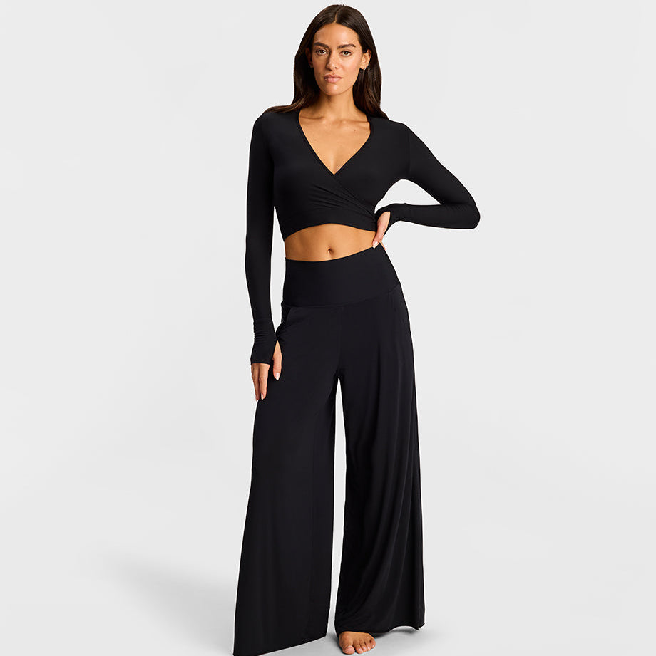 A woman modeling a black two-piece loungewear set consisting of a long-sleeve wrap-style crop top and matching high-waisted wide-leg pants, photographed against a light gray studio background.