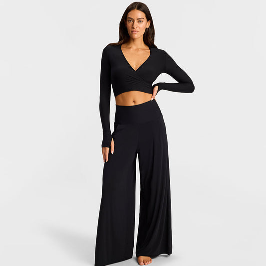 A woman modeling a black two-piece loungewear set consisting of a long-sleeve wrap-style crop top and matching high-waisted wide-leg pants, photographed against a light gray studio background.