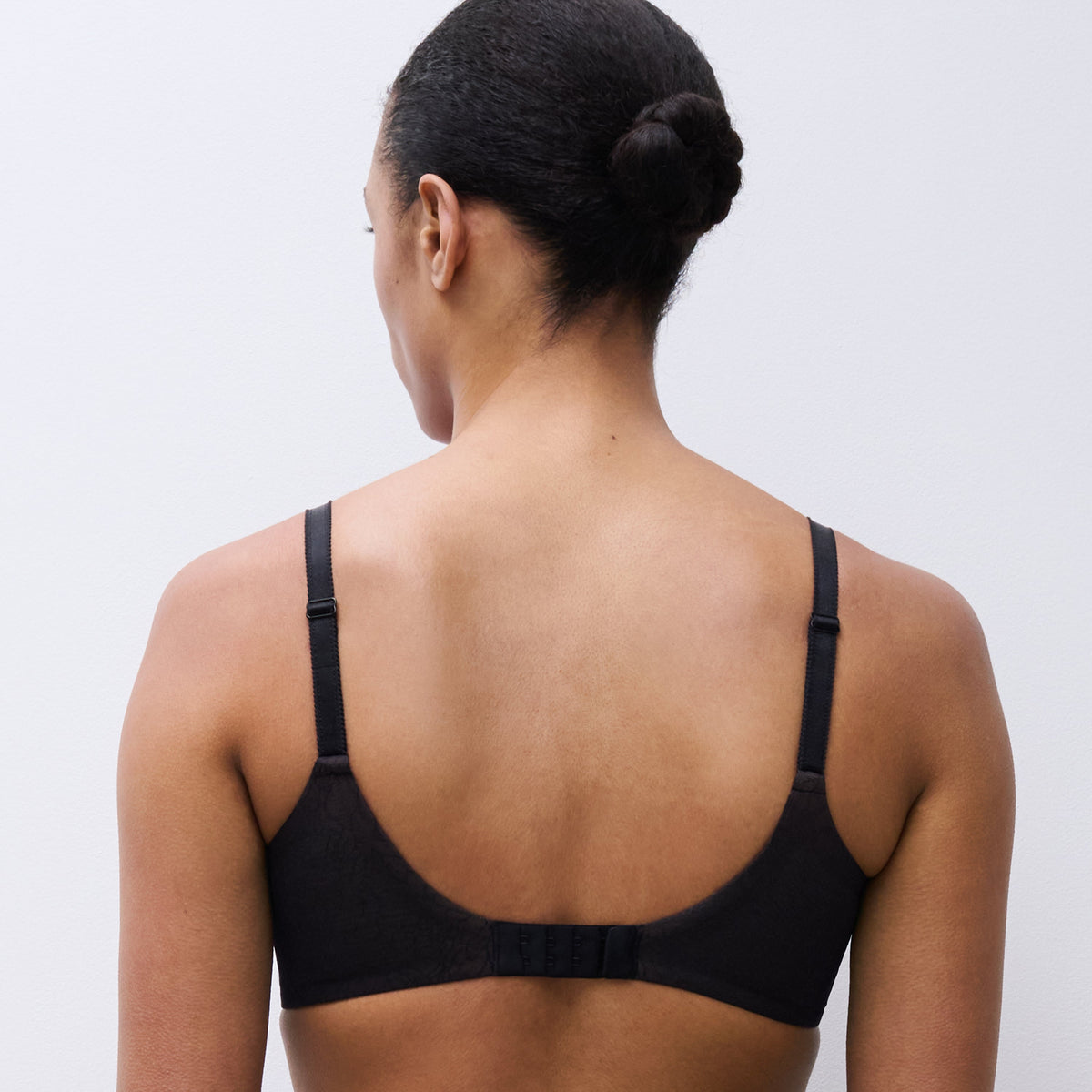A woman viewed from behind wearing a black bra and black underwear against a plain light gray studio background.