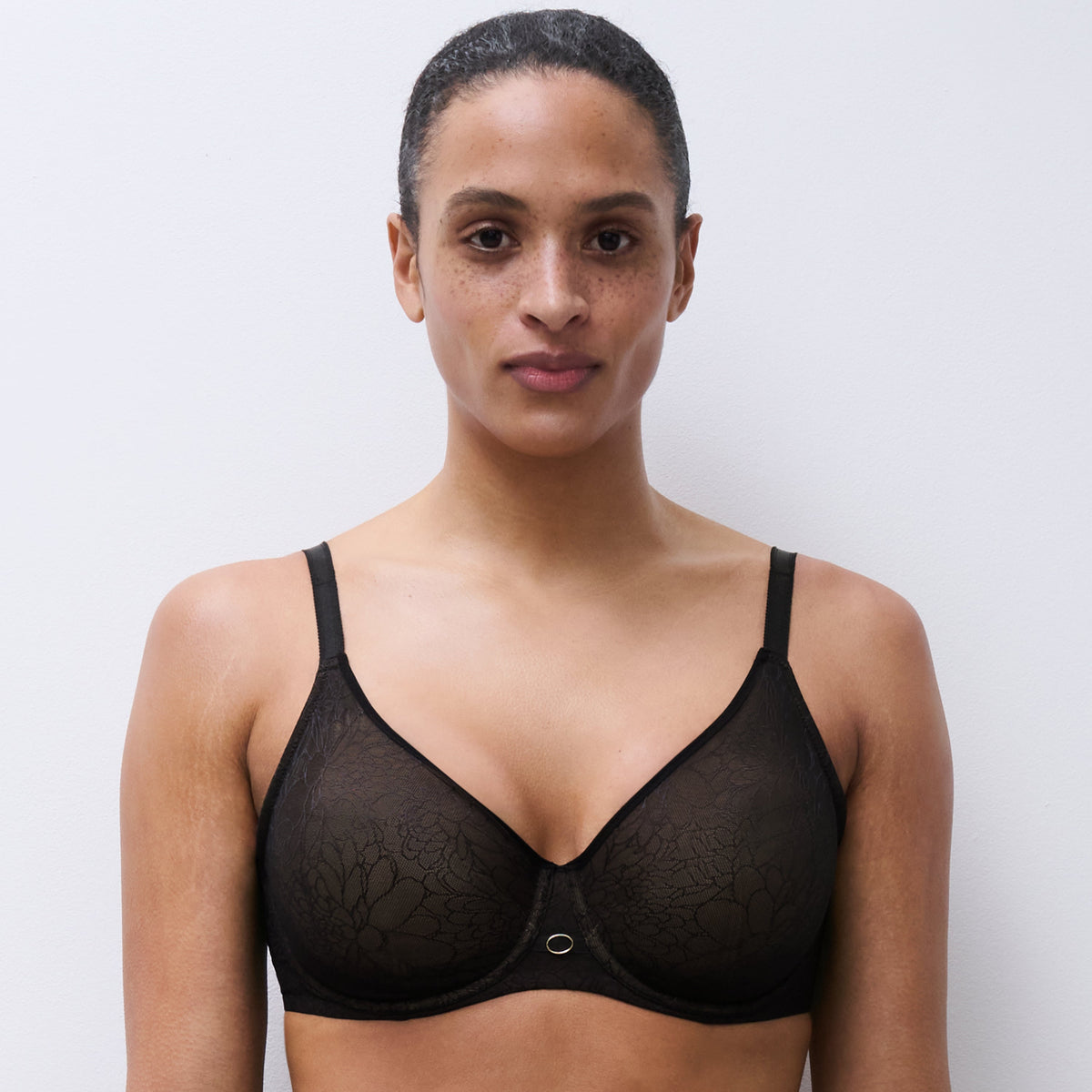 A woman modeling a black sheer lace underwire bra with thin shoulder straps and a small center button, shown against a plain light gray studio background.