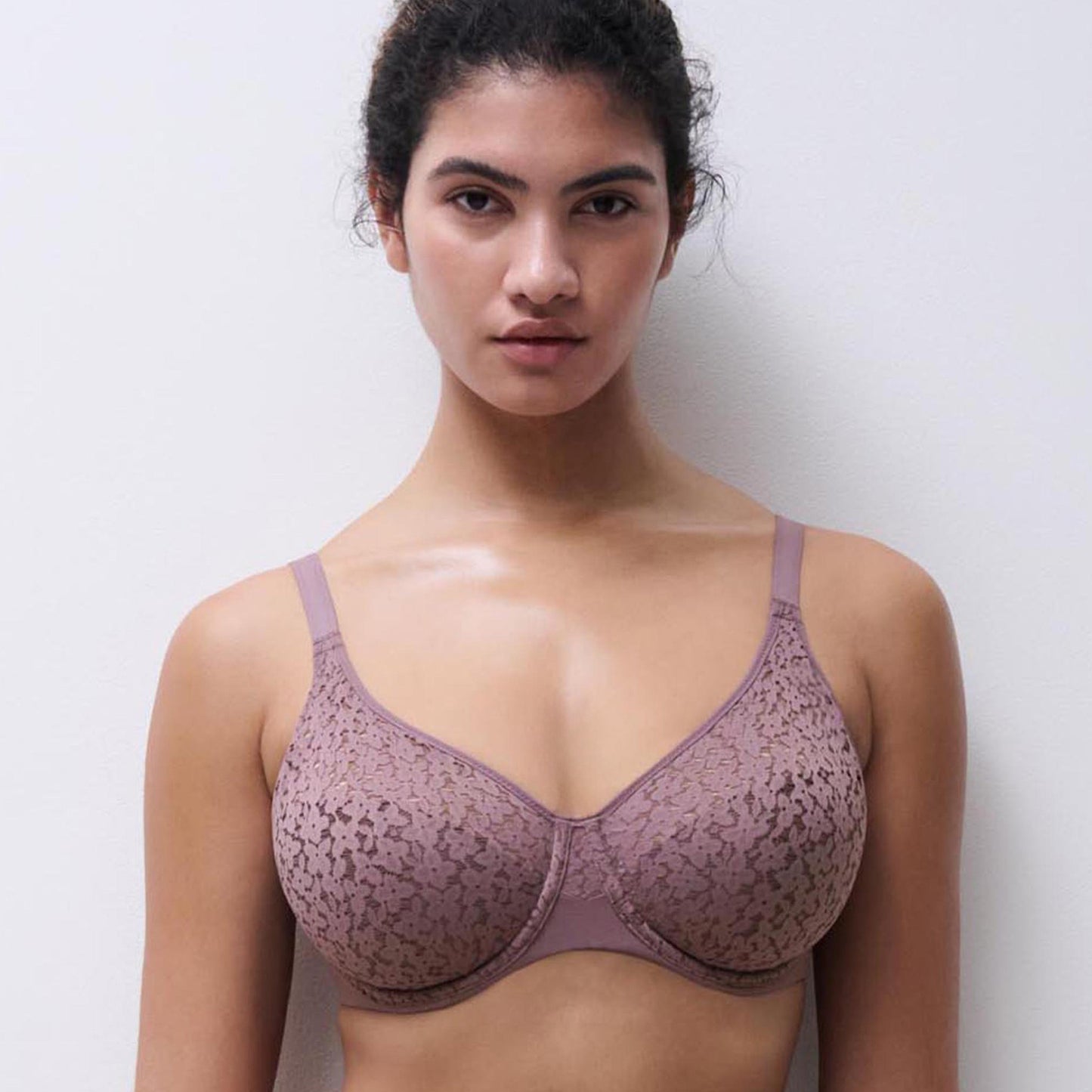 A woman modeling a dusty mauve lace bra against a plain light gray wall.