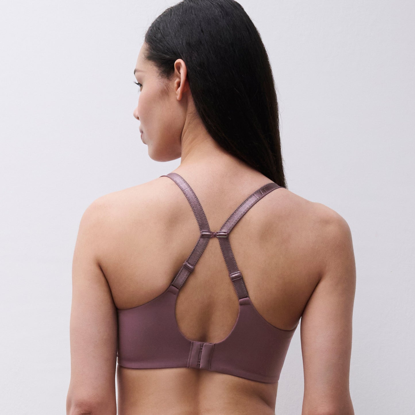 A woman shown from the back wearing a dusty mauve sports bra with crisscross shoulder straps and a small center tie detail, against a plain light gray studio background.