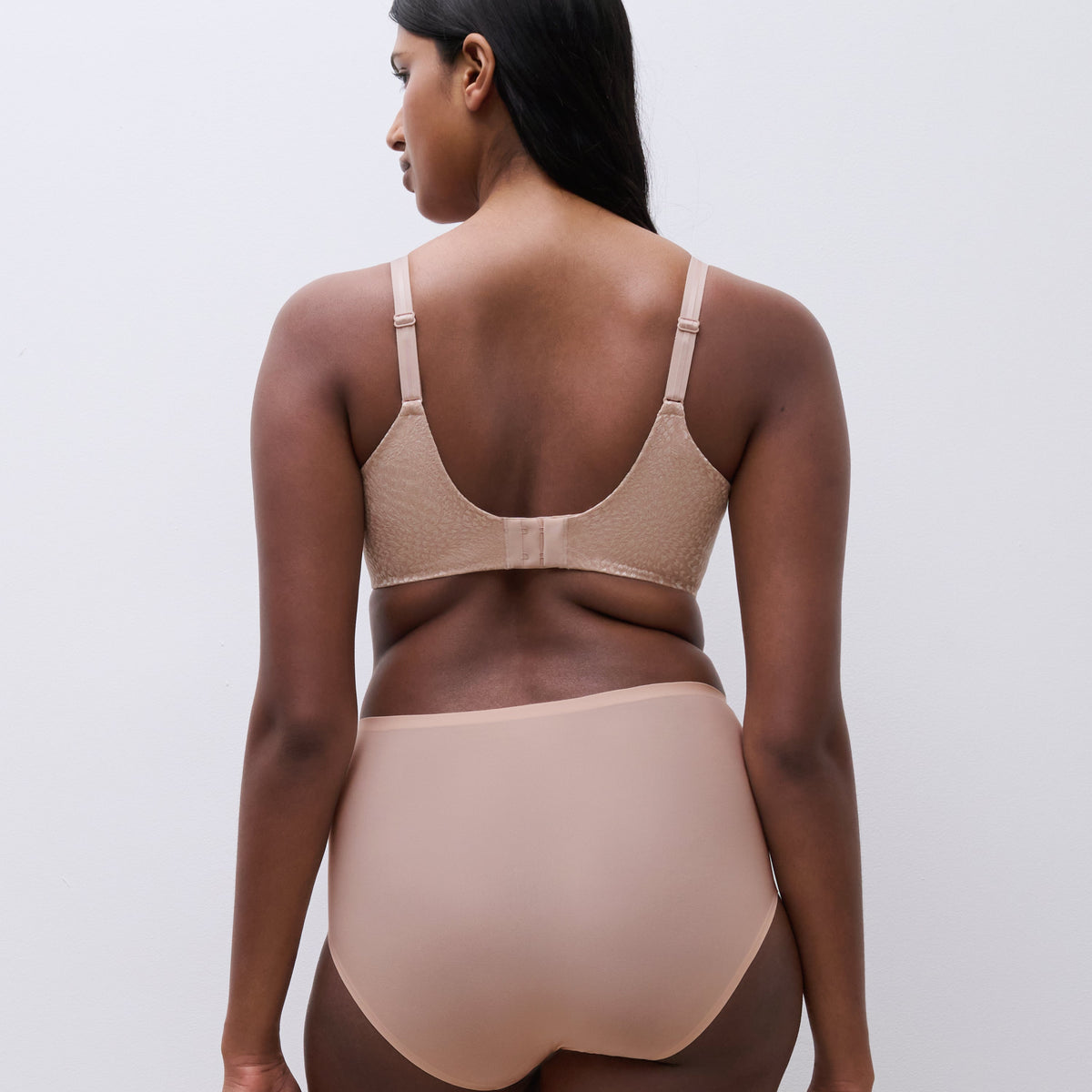A woman shown from the back wearing a beige bra and matching high-waisted shapewear briefs against a plain light gray studio background.