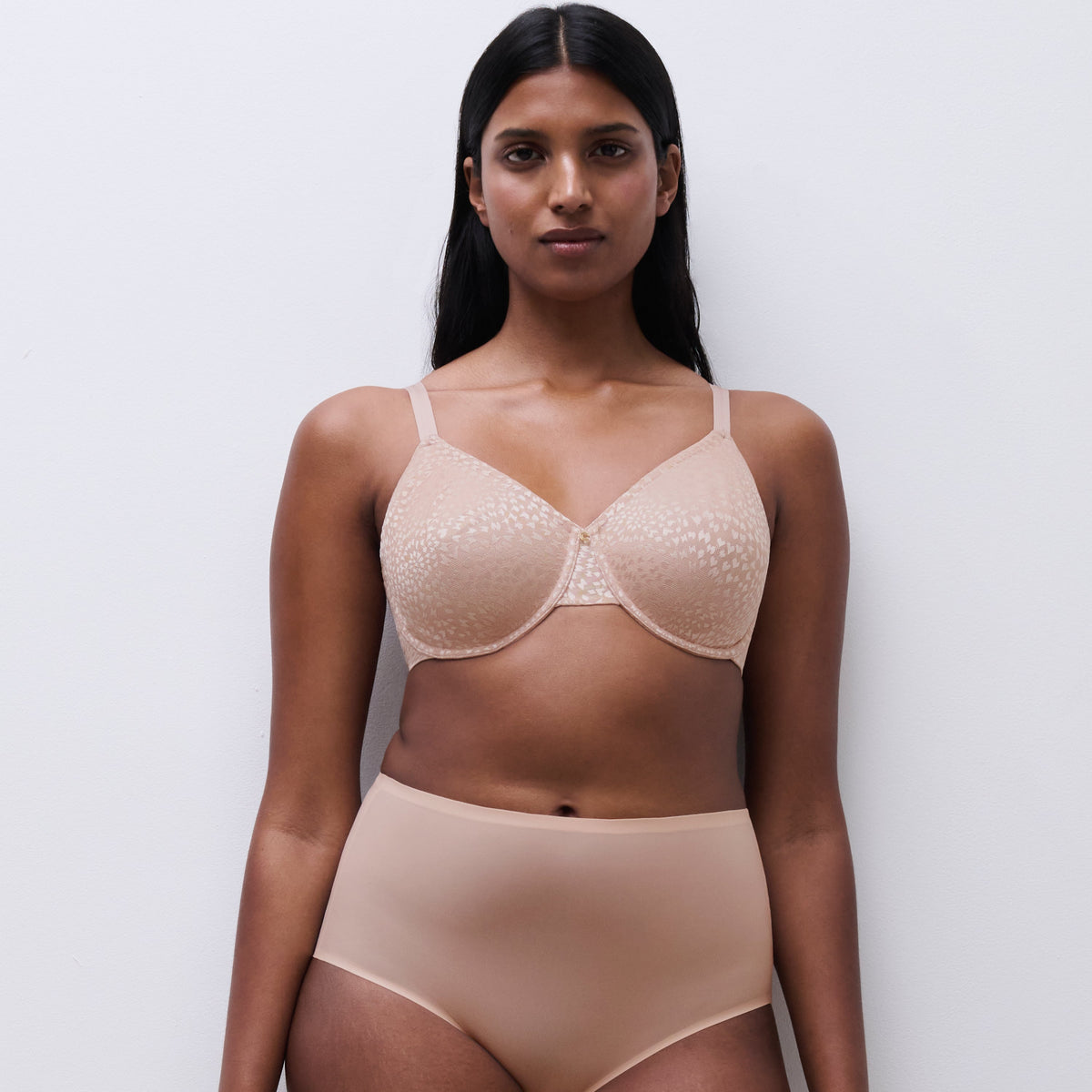 A woman modeling a matching beige lingerie set consisting of a sheer lace underwire bra and high-waisted briefs, standing against a plain light gray background.