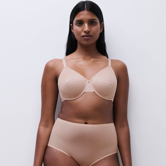 A woman modeling a matching beige lingerie set consisting of a sheer lace underwire bra and high-waisted briefs, standing against a plain light gray background.