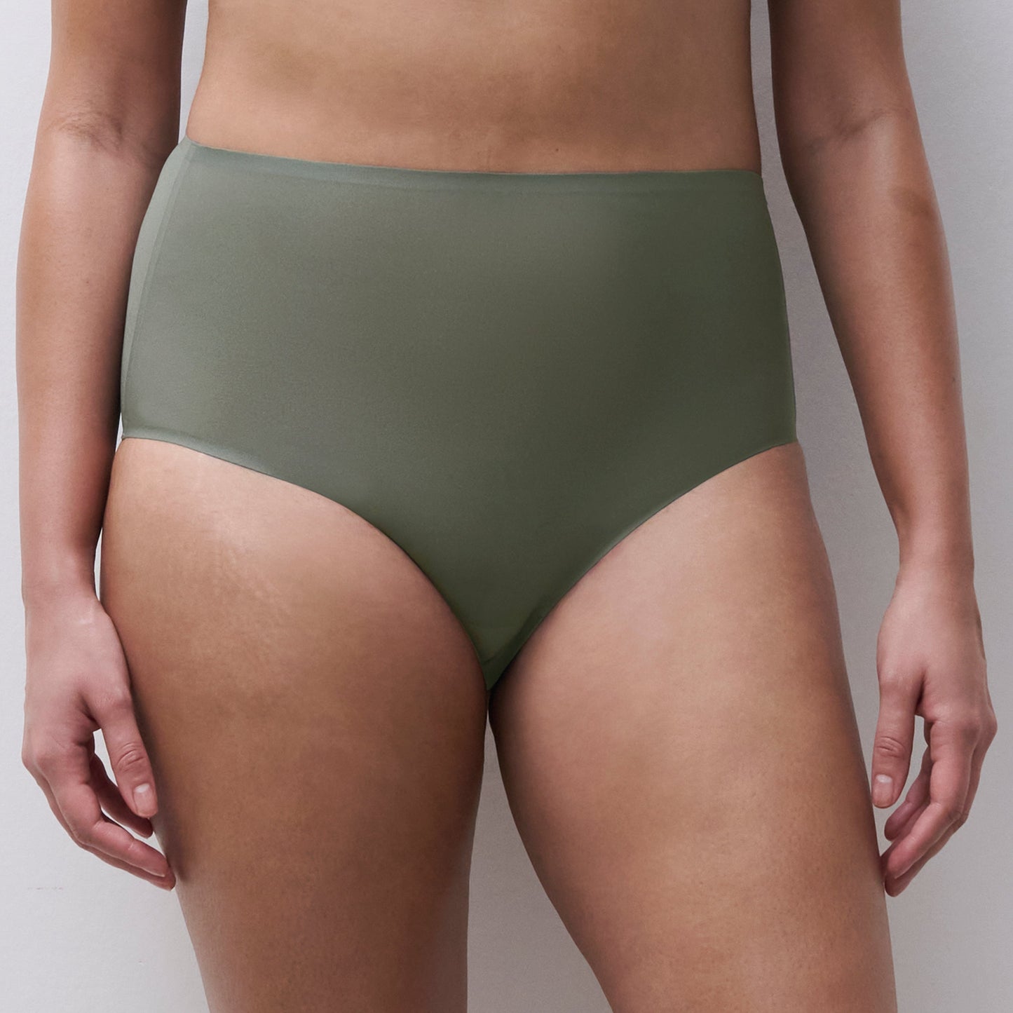 A close-up of a model wearing high-waisted, solid olive green bikini or underwear bottoms against a plain light gray background.