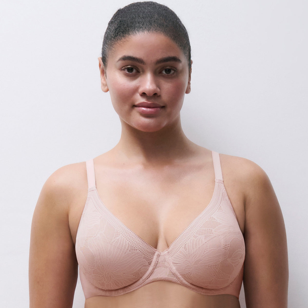 A woman modeling a light beige underwire bra with a subtle floral lace pattern, set against a plain light gray background.