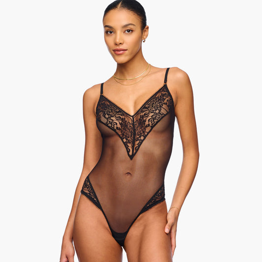 A model wearing a sheer black lace bodysuit with thin straps and a lace-trimmed V-neckline, photographed against a plain light gray studio background.