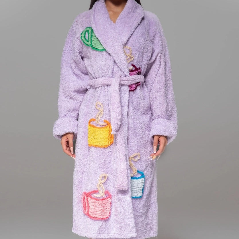 A woman modeling a lavender purple terry cloth bathrobe with embroidered colorful hot drink cups on the front, set against a solid gray studio background.